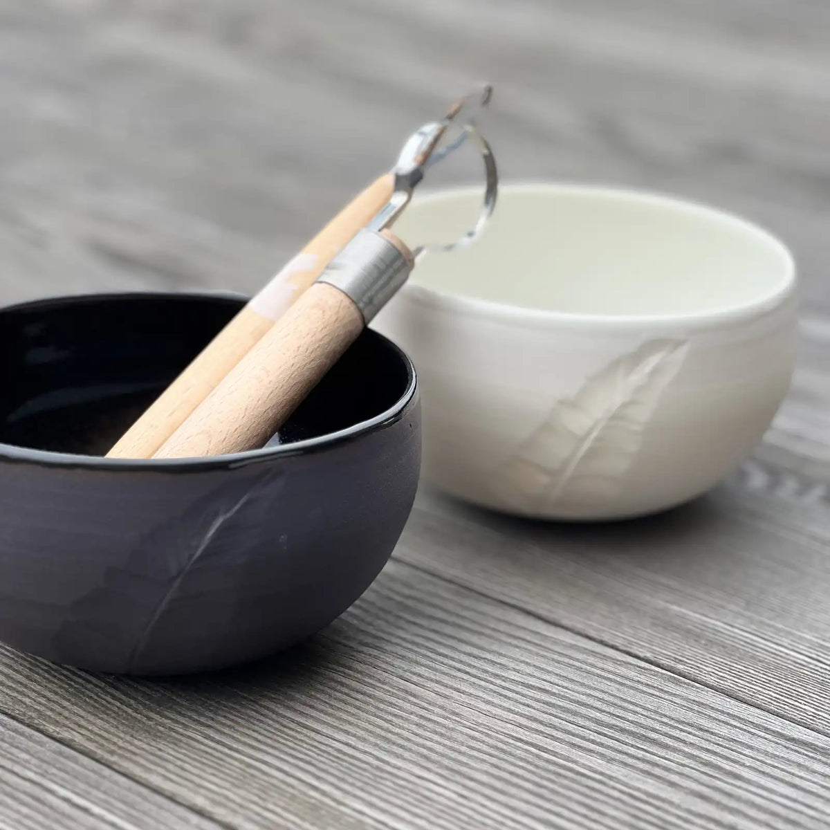 ensemble de bols noir et blanc gravés d'une plume. Deux outils de poterie y sont déposés.